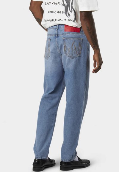 Jeans in denim azzurro con vestibilità comoda, con ricami a fiamma sulle tasche posteriori, indossati con una maglietta grafica bianca e scarpe nere.