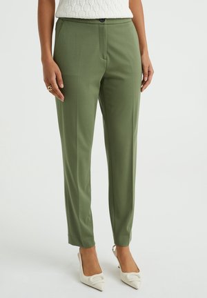 Žena nosící olivově zelené slim-fit kalhoty s předním skladem, bílý pletený top a bílé špičaté podpatky, stojící na jednobarevném pozadí.