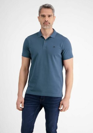 Mann mit salz-und-pfefferfarbenem Haar, der ein tealfarbenes Polohemd mit kurzen Ärmeln und dunkelblaue Jeans trägt und vor einem schlichten weißen Hintergrund steht.