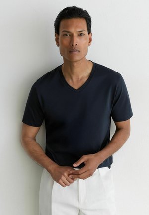 Man in een zwart V-hals T-shirt en witte broek, staand tegen een effen witte muur, handen voor zich geklemd.