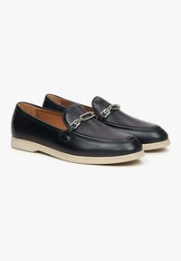 Zwarte leren loafers met een ronde neus, afgewerkt met een zilveren kettingdetail, rustend op een lichtgekleurde rubberen zool. Interieur is tan.