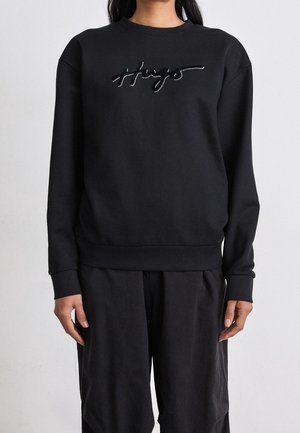 Person wearing a black sweatshirt with a cursive logo on the chest and matching black pants, standing against a plain light background.