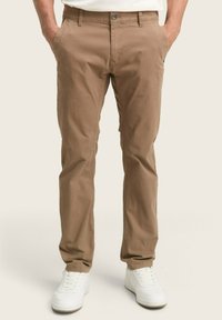 Pantaloni slim-fit, de culoare maro deschis, cu o textură netedă, închidere cu nasture și buzunare laterale, asortați cu adidași albi.