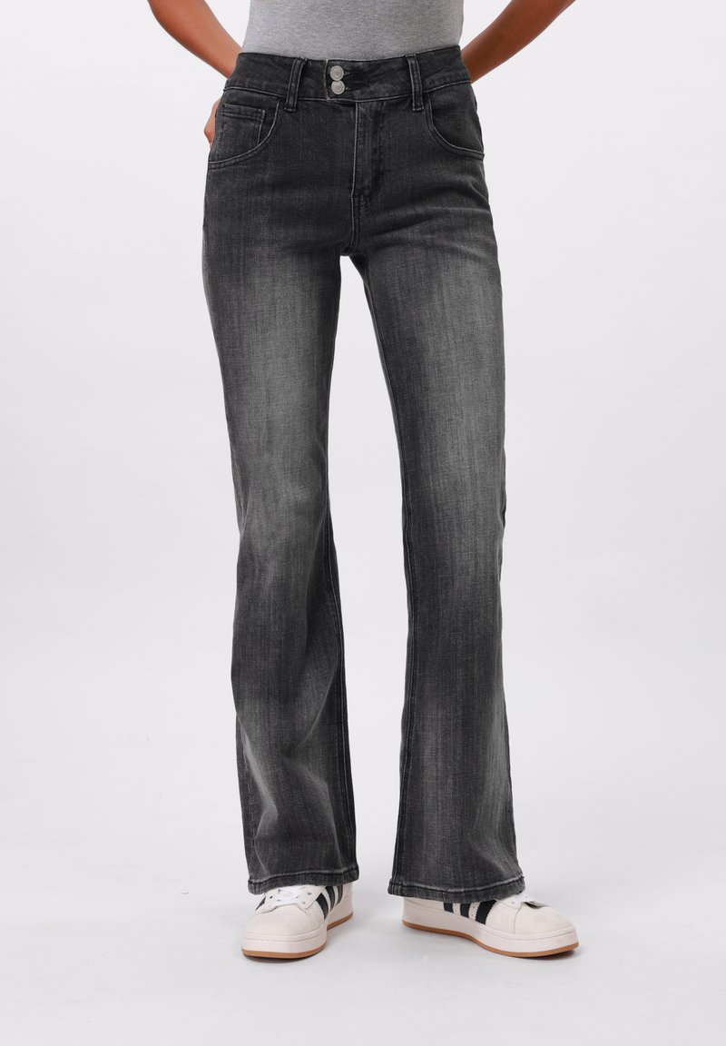 Personne portant un jean bootcut noir délavé et des baskets blanches à rayures noires, debout les mains dans les poches.