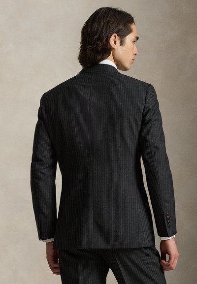 Άνδρας με σκούρα μαλλιά μέχρι τους ώμους, ντυμένος με σκούρο κοστούμι με λεπτές ρίγες και λευκό πουκάμισο, που στέκεται με την πλάτη στην κάμερα μπροστά σε ένα απλό φόντο.