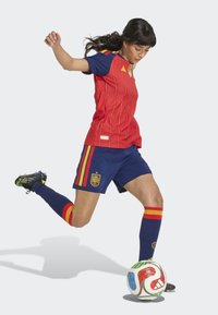 Red soccer jersey with blue accents, featuring vertical stripes and logo; blue shorts with red and yellow details; black cleats. Kicking a soccer ball.