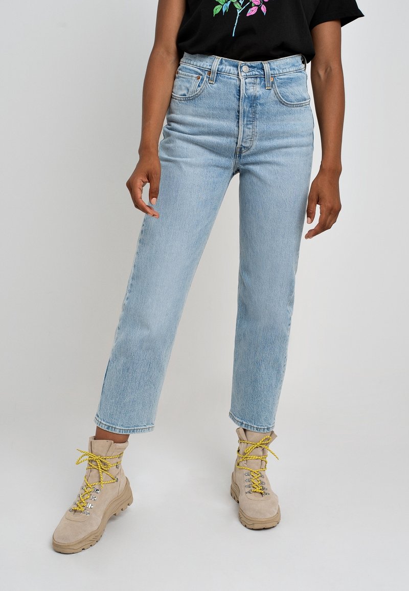 Personne portant un jean taille haute bleu clair et des bottes beiges avec des lacets jaunes, debout devant un fond gris uni.