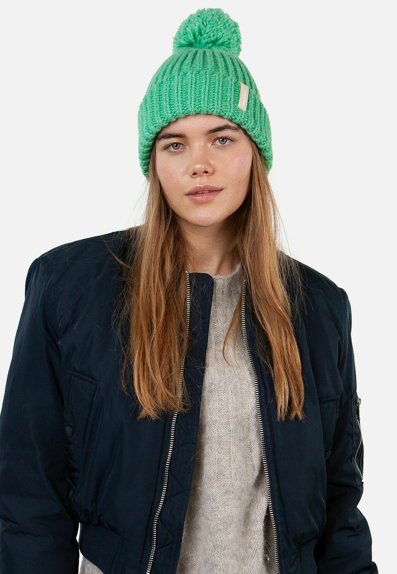 Green knitted beanie featuring a top pom-pom, ribbed texture, and a small label. Worn with a dark bomber jacket and light sweater.