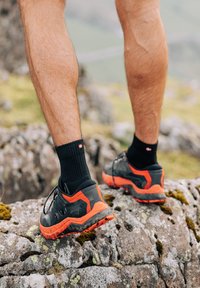 Schwarze und orange Trail-Schuhe mit strukturierten Sohlen und gepolsterten Kragen, getragen mit schwarzen Knöchelsocken, stehen auf unebenen Felsen.