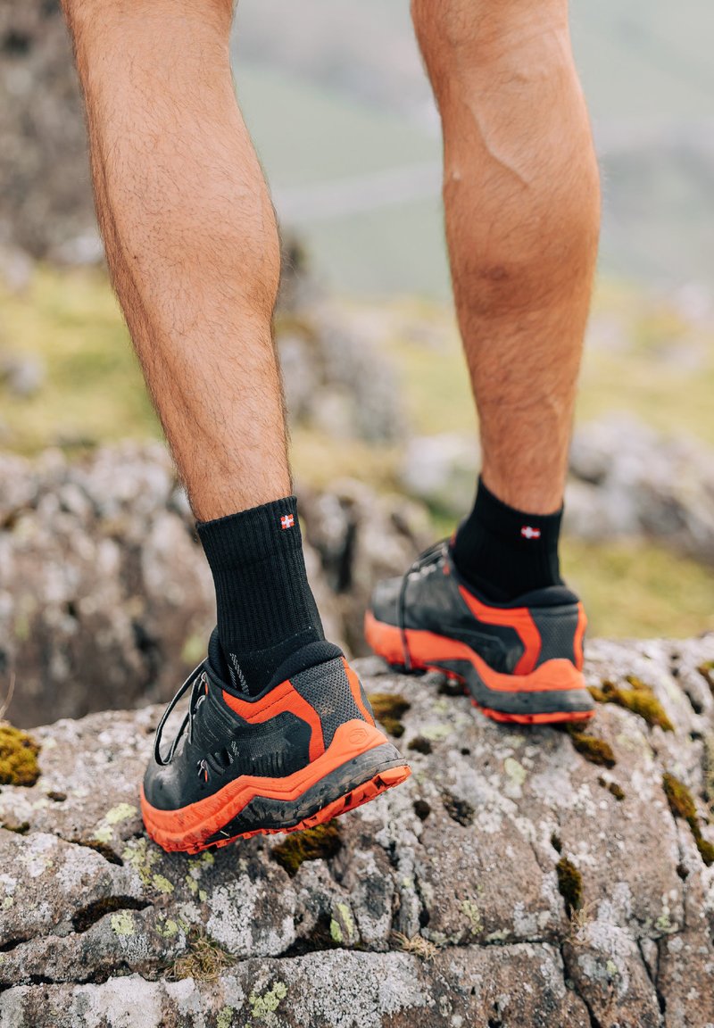 Schwarze und orange Trail-Schuhe mit strukturierten Sohlen und gepolsterten Kragen, getragen mit schwarzen Knöchelsocken, stehen auf unebenen Felsen.