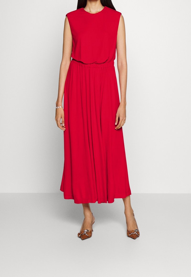 Czerwona, bezrękawnikowa maxi sukienka z okrągłym dekoltem, marszczoną talią i zwiewnym materiałem. Modelka zestawiła ją z brązowymi szpilkami o spiczastym nosku.