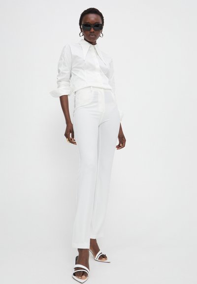 Chemise blanche ajustée avec un grand col, associée à un pantalon blanc sur mesure. Des lunettes de soleil noires et des talons blancs à brides complètent la tenue.