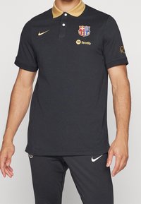 Czarny polo z złotym kołnierzykiem, z herbem FC Barcelona i logo Spotify. Wykonany z gładkiego materiału, z krótkimi rękawami.