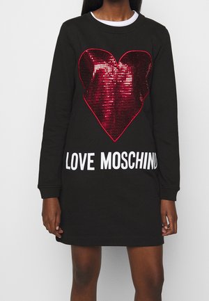 Czarna sukienka z długim rękawem z dużym czerwonym cekinowym sercem i białym napisem "LOVE MOSCHINO" z przodu, noszona przez stojącą osobę.