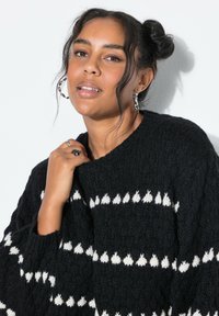 Giovane donna con i capelli scuri raccolti in un bun, che indossa grandi orecchini a cerchio e un maglione nero a maglia con motivi bianchi, in posa contro un muro bianco.