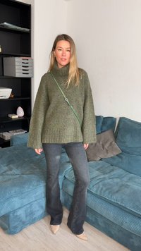 Liels olīvu zaļš adīts džemperis ar augstu apkakli, kopā ar platām tumšām džinsu biksēm un bēšām smailām kurpēm. Zaļa soma pār plecu.