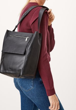 Woman wearing burgundy sweater and blue jeans carrying large black leather shoulder bag with front zip pocket.