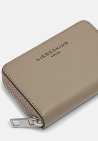 Beżowy skórzany portfel z zamkiem błyskawicznym, o gładkiej fakturze i wytłoczonym nazwiskiem marki "LIEBESKIND BERLIN" z przodu. Srebrne ozdoby metalowe.