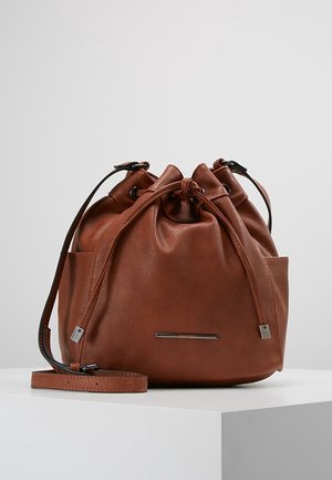 Sac seau en cuir marron avec cordon de serrage, bandoulière réglable, plaque de marque en métal et deux poches latérales extérieures, posé sur une surface blanche.