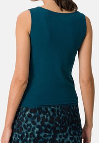 Bezrękawnik w kolorze teal, wykonany z miękkiej tkaniny. Posiada okrągły dekolt i gładką fakturę, zestawiony ze spódnicą w panterkę w odcieniach niebieskiego i czarnego.