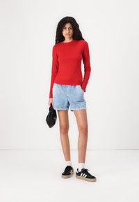 Top roșu cu mâneci lungi, ajustat, asortat cu pantaloni scurți din denim albastru deschis. Modelul poartă adidași negri cu sosete albe și ține o geantă neagră.
