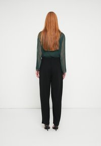 Black high-waisted trousers paired with a sheer green long-sleeve blouse. Long red hair falls down the back, creating texture contrast.