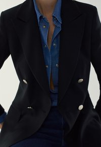 Blazer noir croisé avec des boutons argentés, superposé à une chemise en denim bleu. Possède des revers crantés et une coupe cintrée.