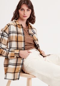 Camisa de cuadros multicolor en amarillo, blanco y negro, hecha de material suave, con un ajuste relajado, llevada sobre una parte superior texturizada marrón y pantalones crema.