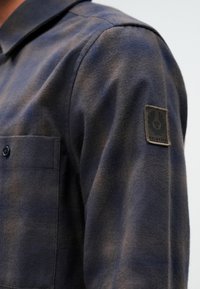 Tumši zila flaneļa krekls ar krūšu kabatu, ar tekstūras audumu un melnu uzlīmi ar "Belstaff" logotipu uz piedurknes.