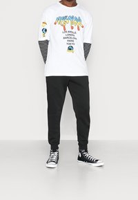 En person som bär en vit långärmad skjorta med färgglad text "New York" och stad namn, svarta randiga ärmar, svarta byxor och svarta högkängor.