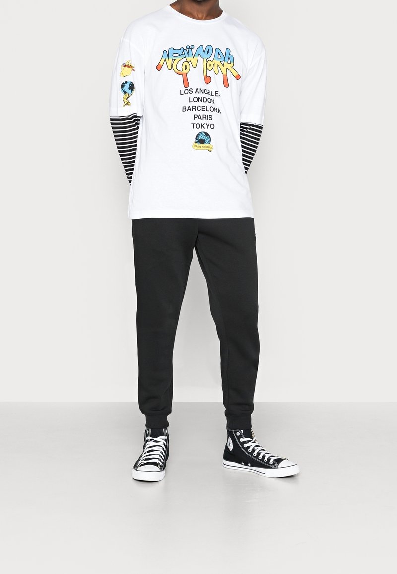 En person som bär en vit långärmad skjorta med färgglad text "New York" och stad namn, svarta randiga ärmar, svarta byxor och svarta högkängor.