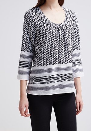 Frau trägt ein schwarz-weiß gemustertes Dreiviertelarm-Top mit Streifen und geometrischen Designs, kombiniert mit schwarzen eng anliegenden Hosen.