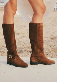 Botine din suede maro, până la genunchi, cu talpă plată, fermoar pe lateral și o curea cu cataramă metalică. Designul prezintă o textură netedă și o siluetă simplă.