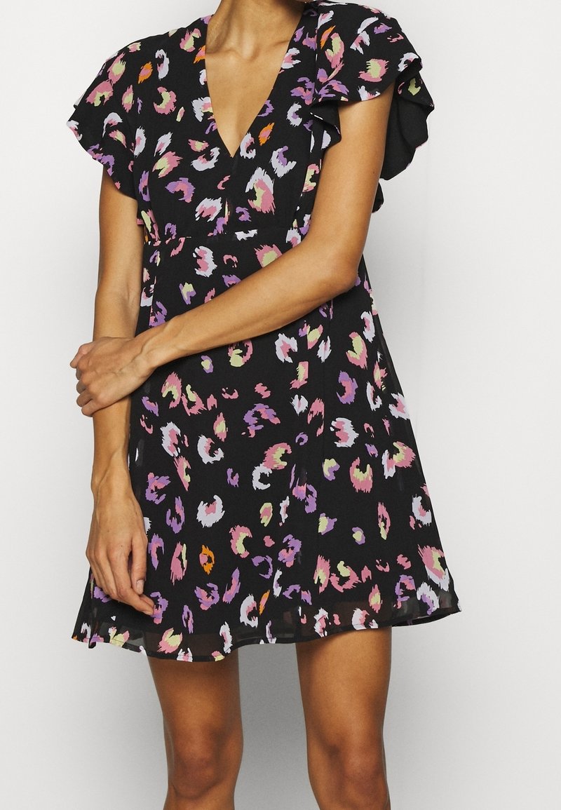 Femme portant une courte robe noire avec un motif floral abstrait coloré et des manches courtes à volants, debout les bras croisés.