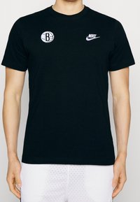 Camiseta de algodón negra con un logo circular blanco de baloncesto de Brooklyn en el lado izquierdo del pecho y un swoosh blanco de Nike en el lado derecho. Mangas cortas.