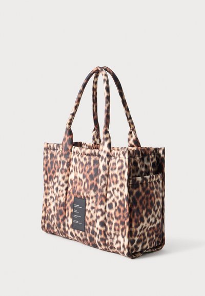 Sac fourre-tout en tissu imprimé léopard avec double poignée et une petite étiquette noire rectangulaire à l'avant sur un fond uni.