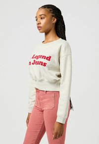Felpa grigia chiaro con ricamo rosso "Legend in Jeans", abbinata a jeans rosa. Presenta un collo rotondo e polsini a costine.
