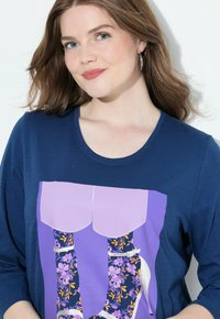 Tmavě modrá košile s dlouhými rukávy, zdobená grafikou květinových bot s fialovými akcenty a stříbrnými podpatky. Viditelná měkká textura látky.