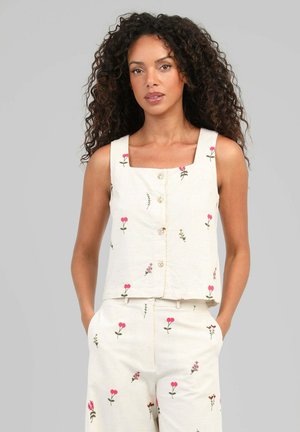 Femme aux cheveux bouclés portant un haut crème sans manches boutonné et un pantalon assorti avec de petits imprimés floraux roses, mains dans les poches, fond gris.