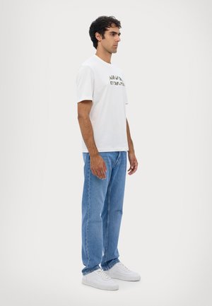 T-shirt bianca in cotone con testo stampato, abbinata a jeans di denim blu chiaro. Modello che indossa sneakers bianche, in posizione laterale.