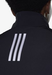 Chaqueta negra con un cuello alto que presenta tres franjas verticales plateadas en la parte posterior. Textura suave, diseño minimalista.