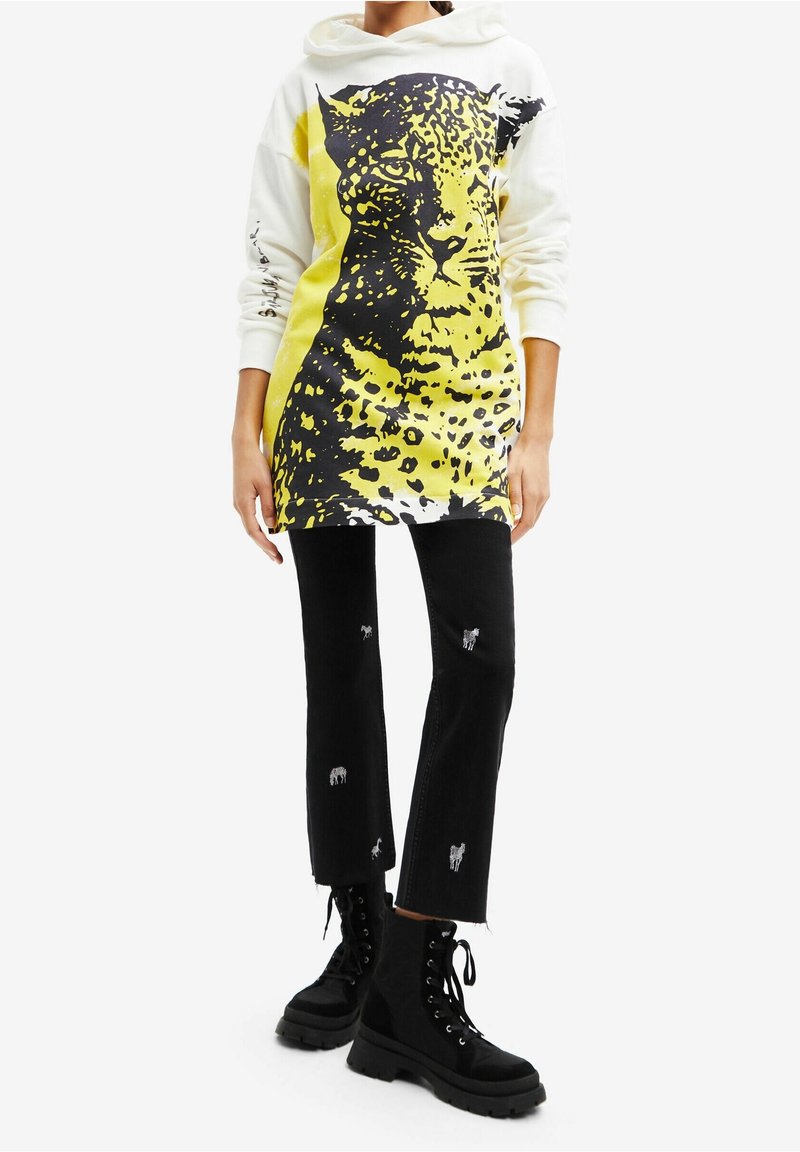Personne portant un sweat à capuche blanc avec un grand motif de visage de léopard jaune et noir, un jean noir avec une petite broderie d'animal, et des bottes noires à lacets.