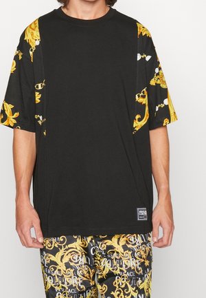 Mężczyzna w czarnej koszulce z żółtym barokowym wzorem na rękawach i pasujących spodenkach z wzorem oraz białym napisem na czarnym tle.