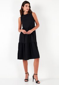 Black sleeveless dress with tiered ruffle design, made of lightweight fabric. The hemline falls below the knee, paired with black high-heeled shoes.