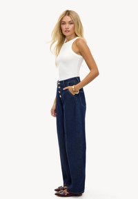 Vaqueros de mezclilla azul oscuro de tiro alto, pierna ancha y corte recto, con botones dorados y bolsillos delanteros, combinados con una camiseta sin mangas blanca.