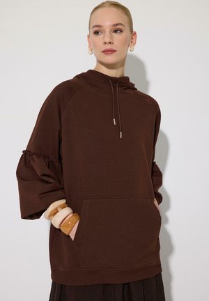 Femme aux cheveux blonds porte un sweat à capuche marron avec des manches à volants et de grands bracelets beiges et marrons, debout devant un mur clair uni.