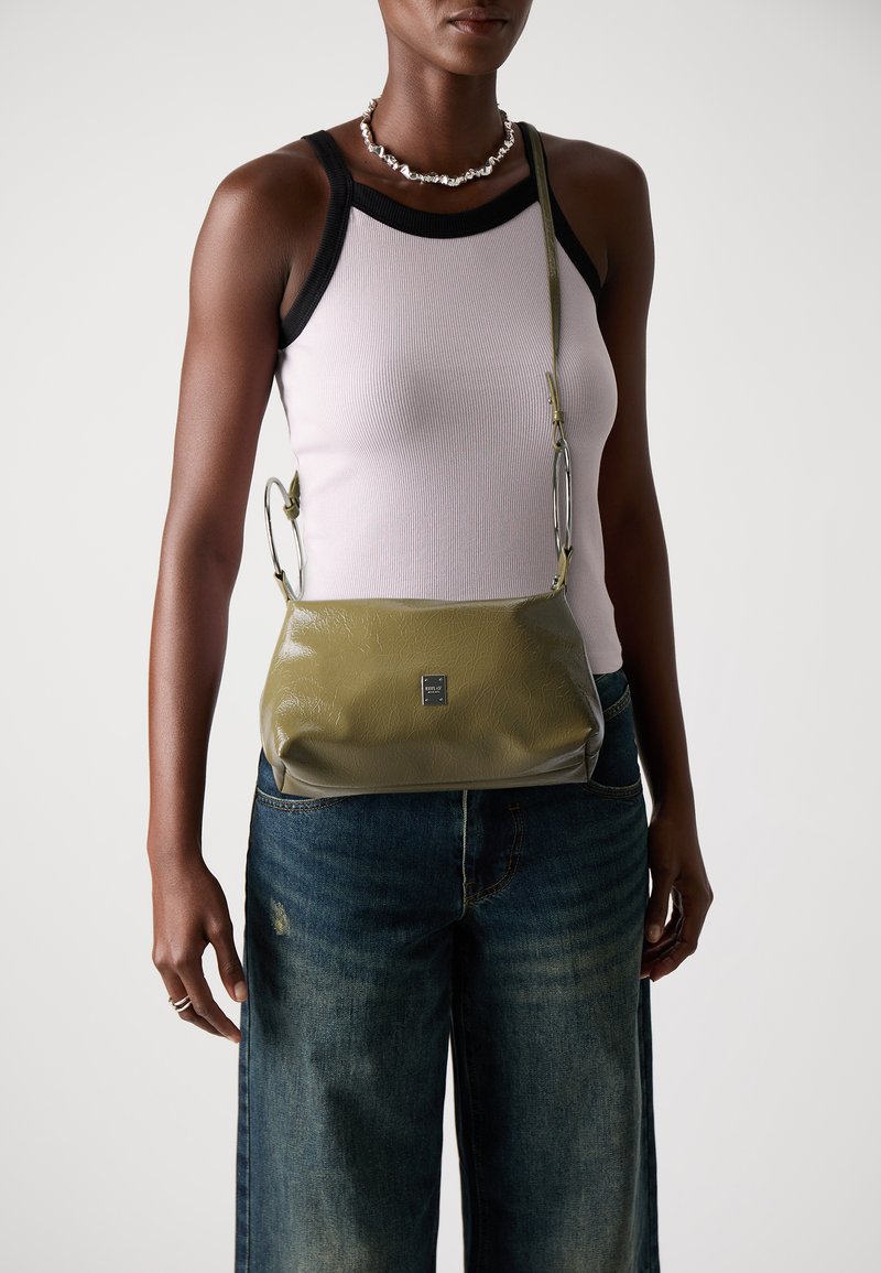 Grønn lærveske med teksturert overflate, med en sølvkjedestropp og en liten logo på forsiden. Bært med denimjeans.