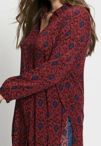 Chemise rouge boutonnée avec un motif orné d'accents bleus. Le tissu est léger, à manches longues, avec des fentes latérales pour faciliter les mouvements.