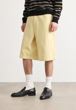 Gule bomullsshortser med løs passform, rett kant og sidelommer, parret med svarte slip-on sko og hvite ribbestrikkede sokker.