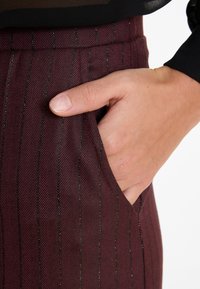 Hand rustend in de zak van bordeauxrood gestreepte broek, met een zwarte top die zichtbaar is bij de taille.